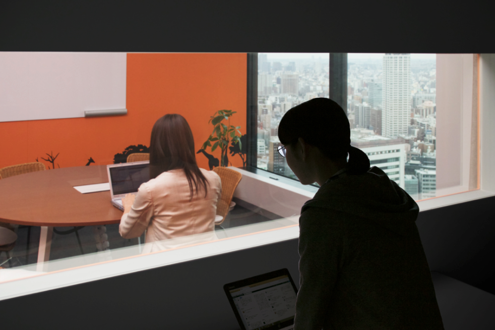 オレンジ色の壁の部屋の中でパソコンを操作する人の様子を別室から見つめる人の後ろ姿の写真。