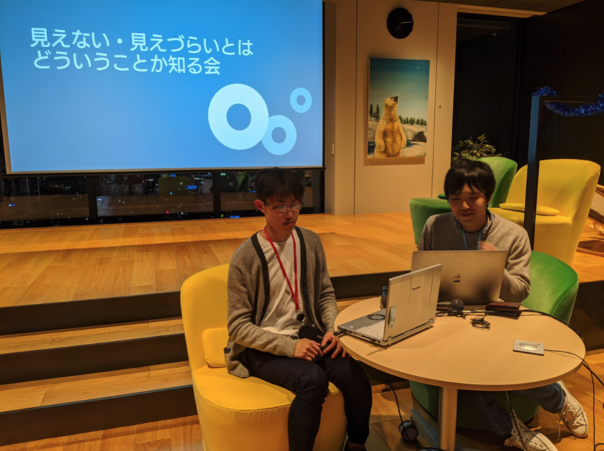 写真：SUGIさんと開催した勉強会「見えない・見えづらいとはどういうことか知る会」の様子。スクリーンを背に、男性2名がパソコンに向かって座っている。
